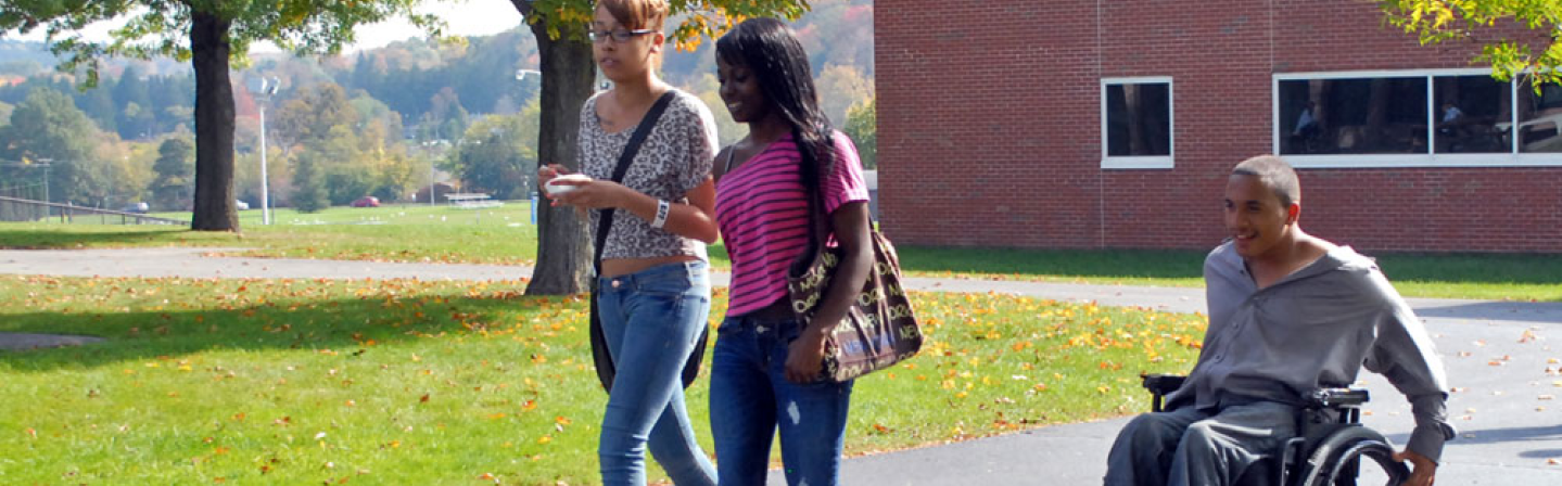 Three students outside on a college campus.  Two of the students are walking and one is traveling with them by wheelchair.